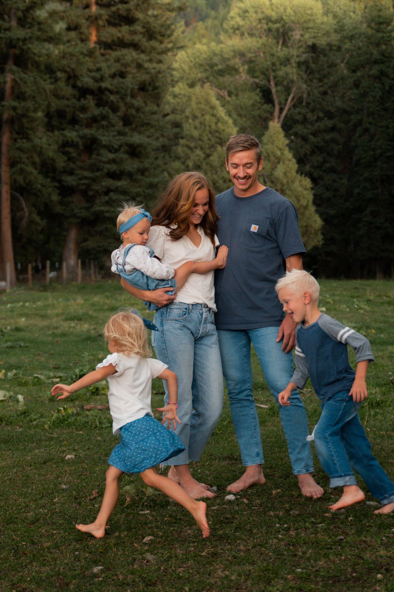 A happy family of five plays barefoot on green grass with tall pine trees in the background at sunset.