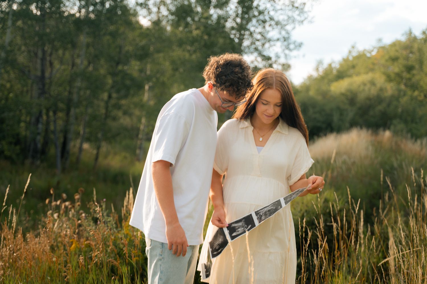 Couple examining ultrasound photos together in a sunny field, expecting parents sharing a tender moment.
