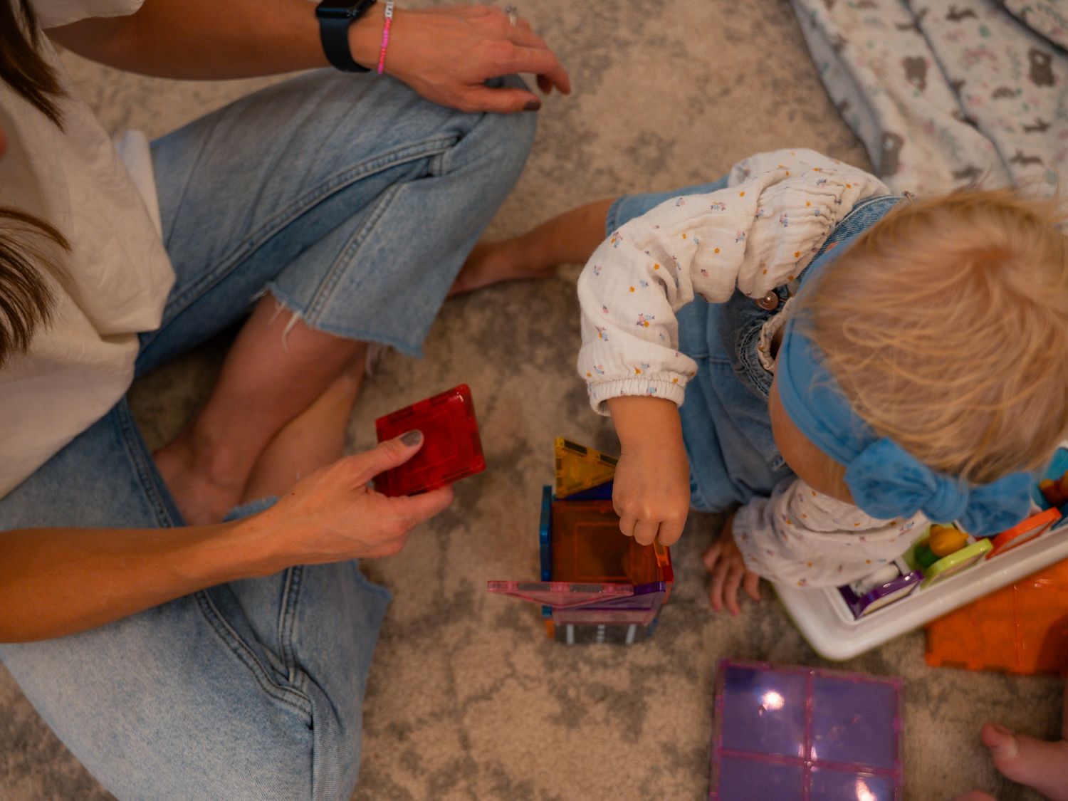Mother sits on carpet playing with colorful toys alongside her young toddler in denim overalls.