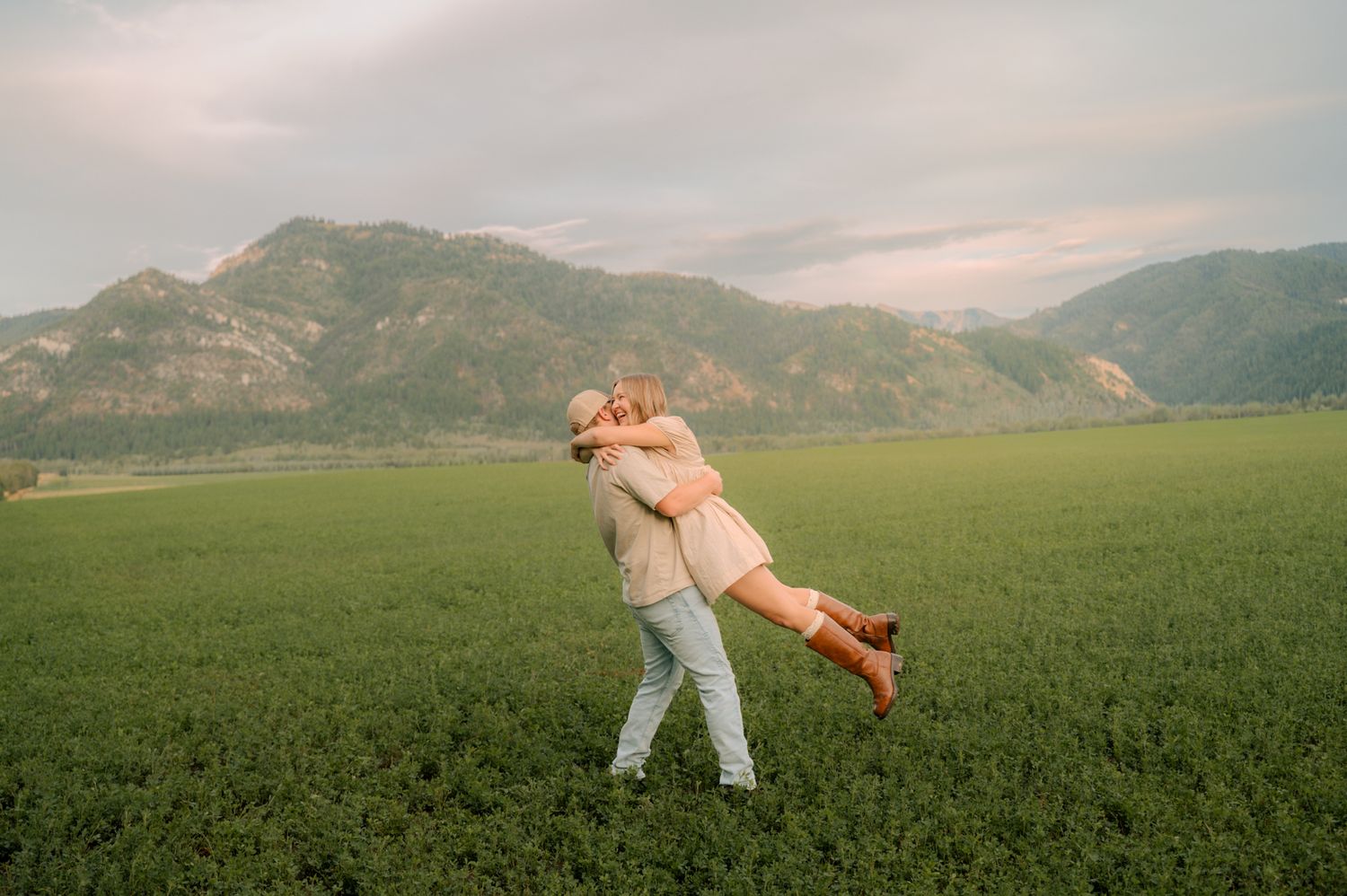 Man lifting woman in joyful embrace in lush green field with mountains.