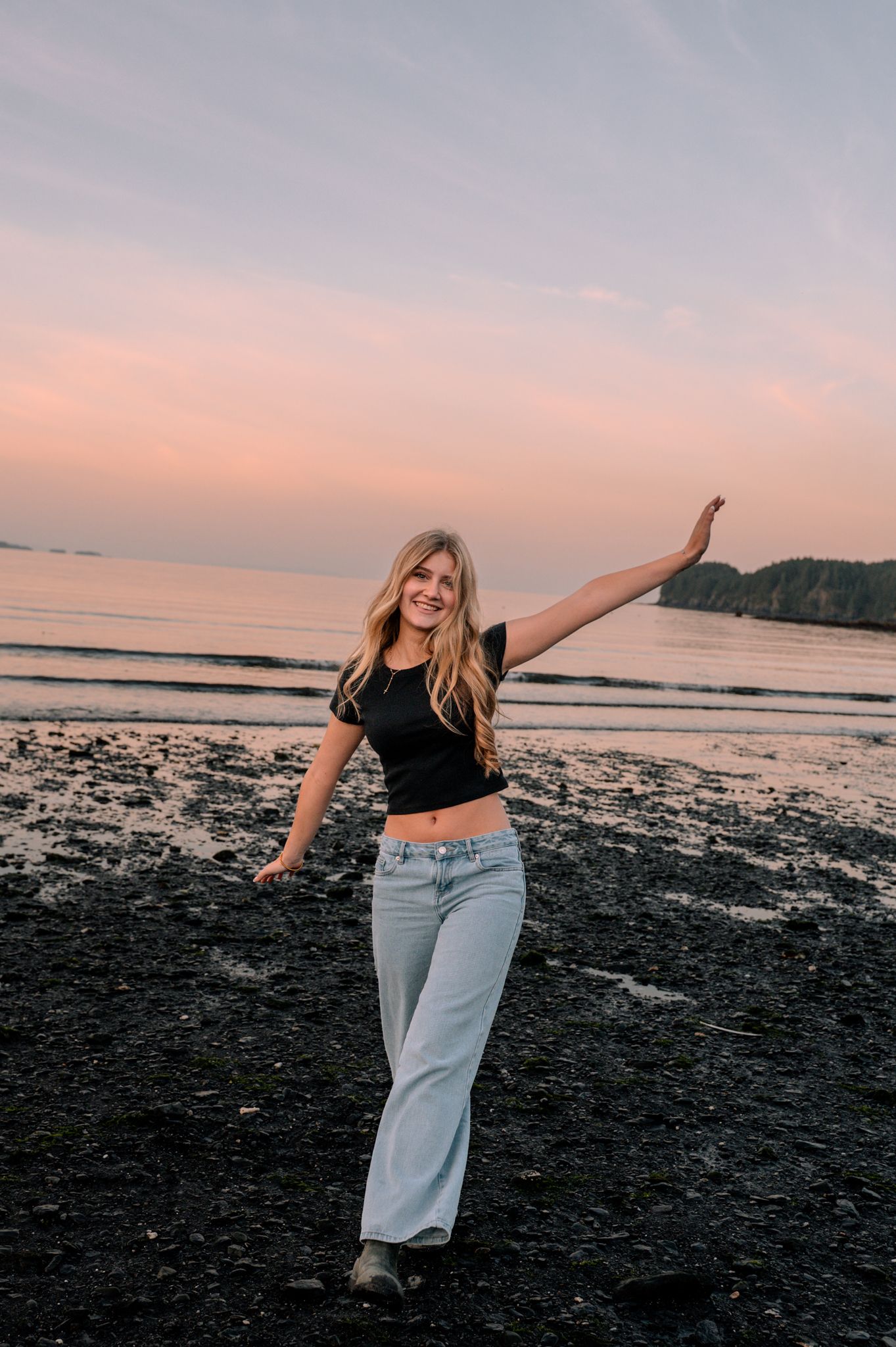 Young woman in black crop top and wide-leg jeans poses on rocky beach at sunset with arm raised.