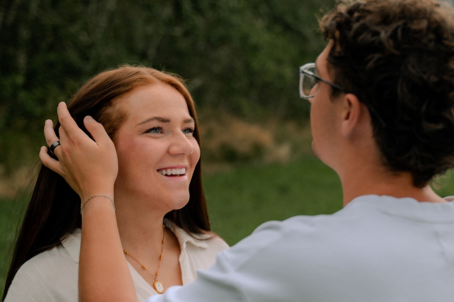 Smiling young woman with brown hair having her hair tucked back by a man in a sunny outdoor setting.