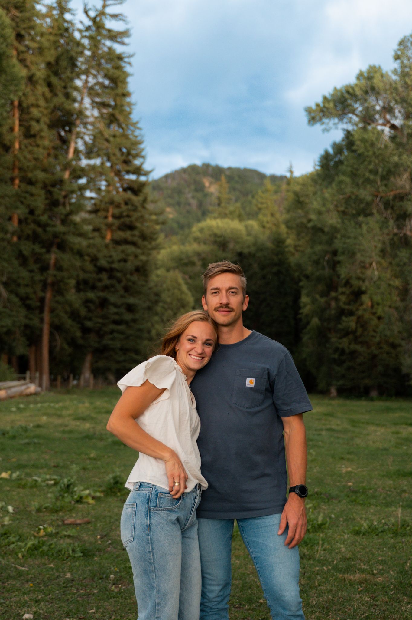 Couple embracing in a mountain forest meadow at golden hour, woman in white top and man in navy Carhartt tee.