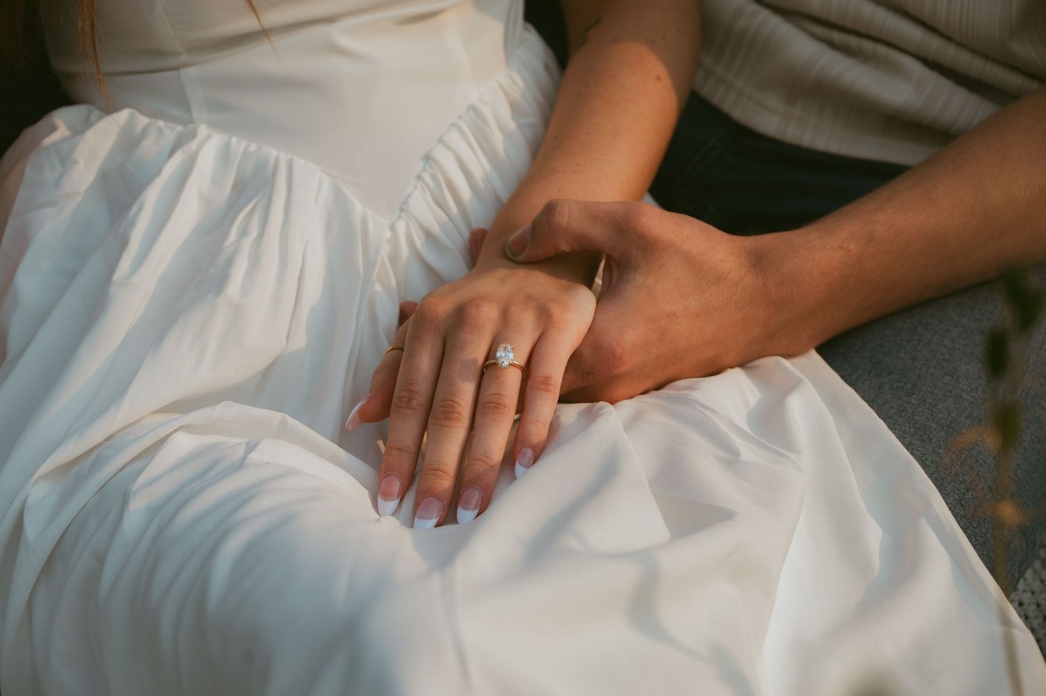 Close-up of a man's hand resting on a woman's hand wearing a diamond engagement ring, her white flowing dress draped softly around them.