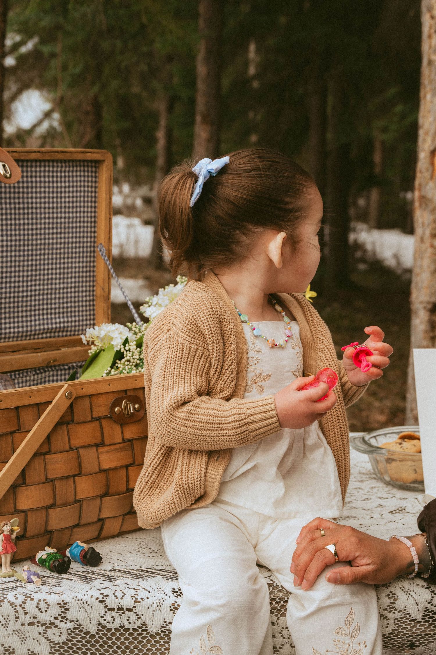 Toddler in a knit cardigan and white overalls sits on a lace tablecloth beside a wicker picnic basket in a forest, holding small red toys.