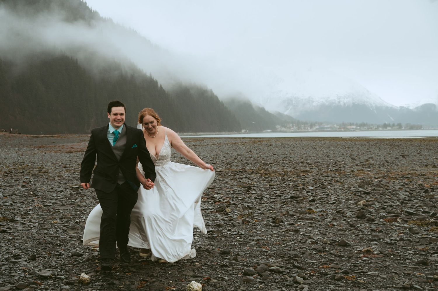 Bride and groom walking hand in hand on a rocky beach with misty mountains in the background on their wedding day.