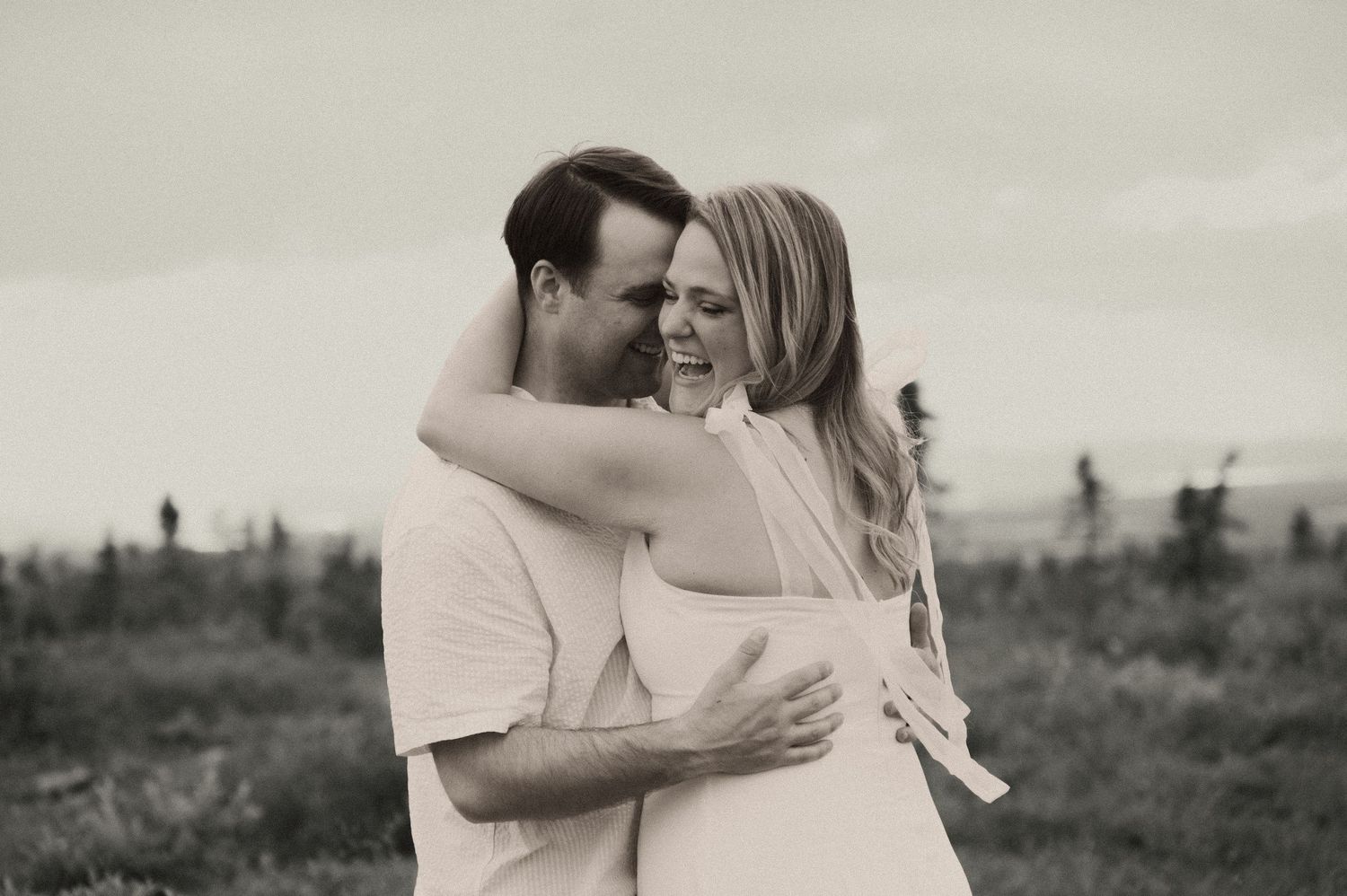Black and white photo of a smiling couple embracing in a grassy field with evergreen trees and a cloudy sky behind them.