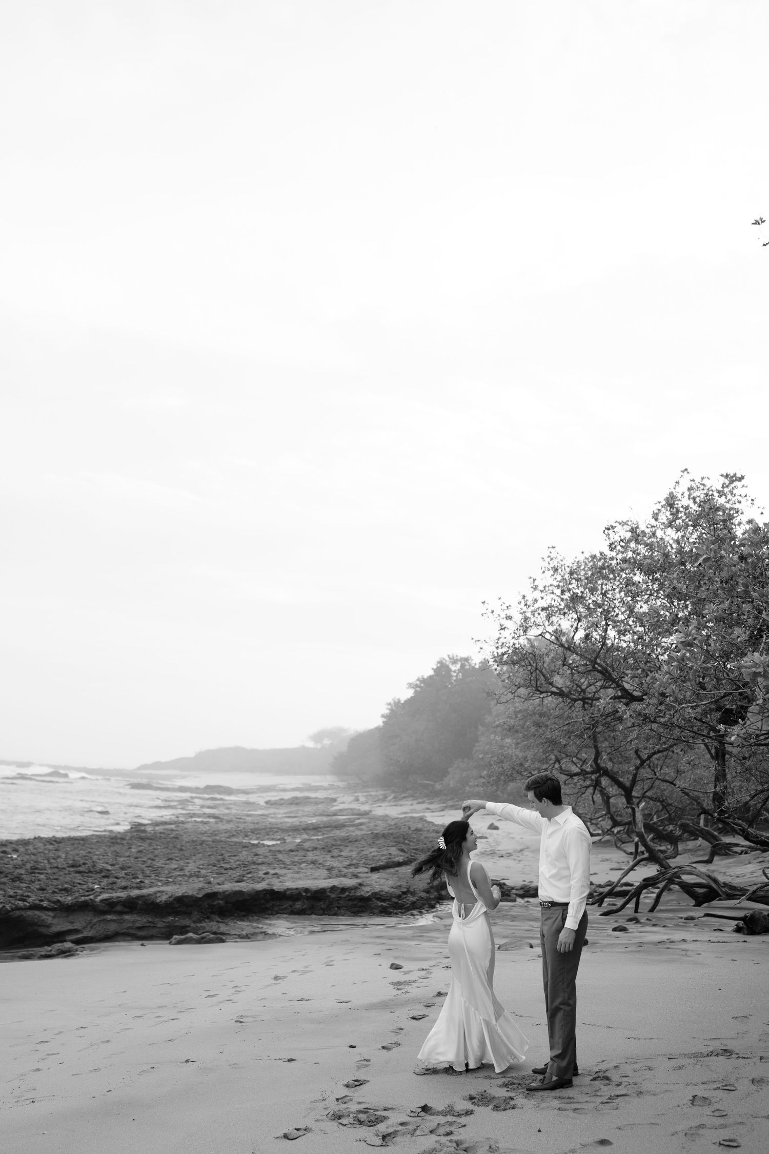 Black and white photo of a couple dancing on a misty beach, the bride in a flowing gown twirling beside rocky shores and windswept trees.
