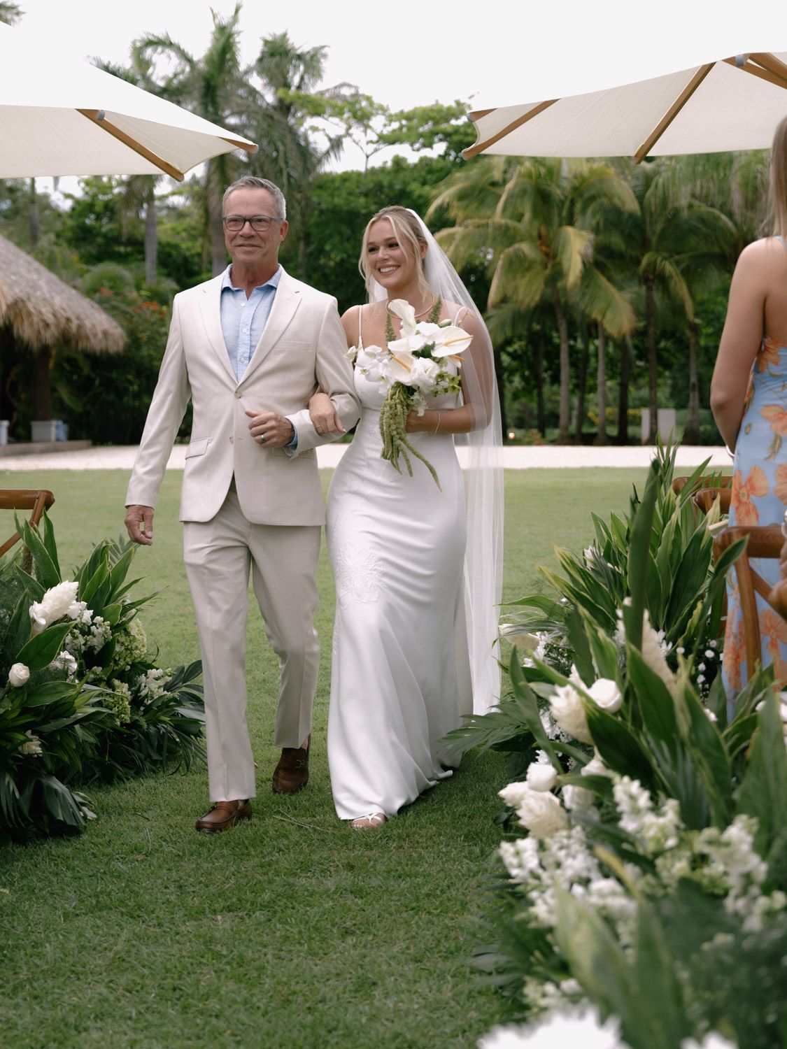 A man in a cream suit walks a bride in a white gown and veil down a grassy outdoor aisle in costa rica