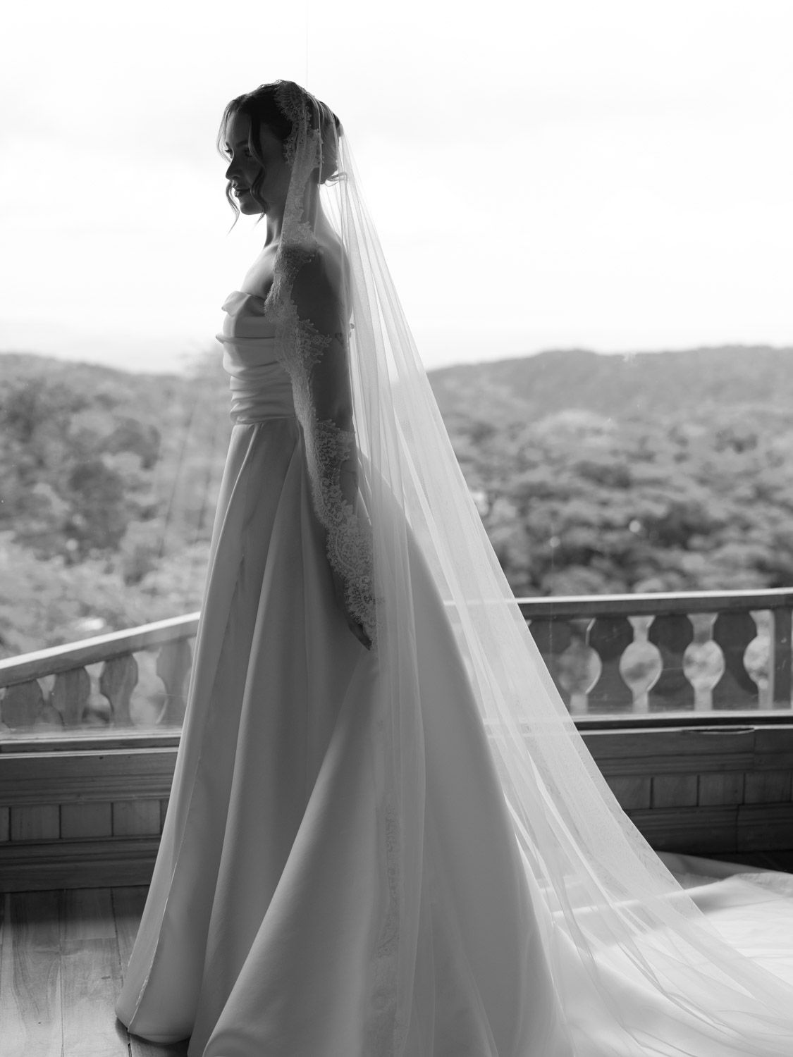 Bride in elegant white wedding gown and long veil stands on balcony overlooking lush greenery in black and white photo.