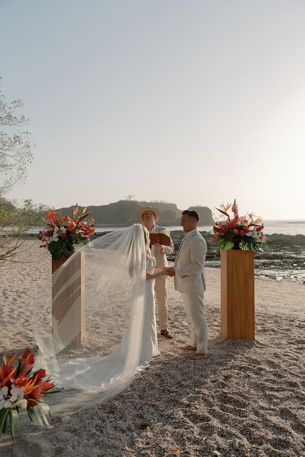 Bride and groom holding hands during a beach wedding ceremony in Costa Rica, framed by tropical flowers in warm afternoon light.