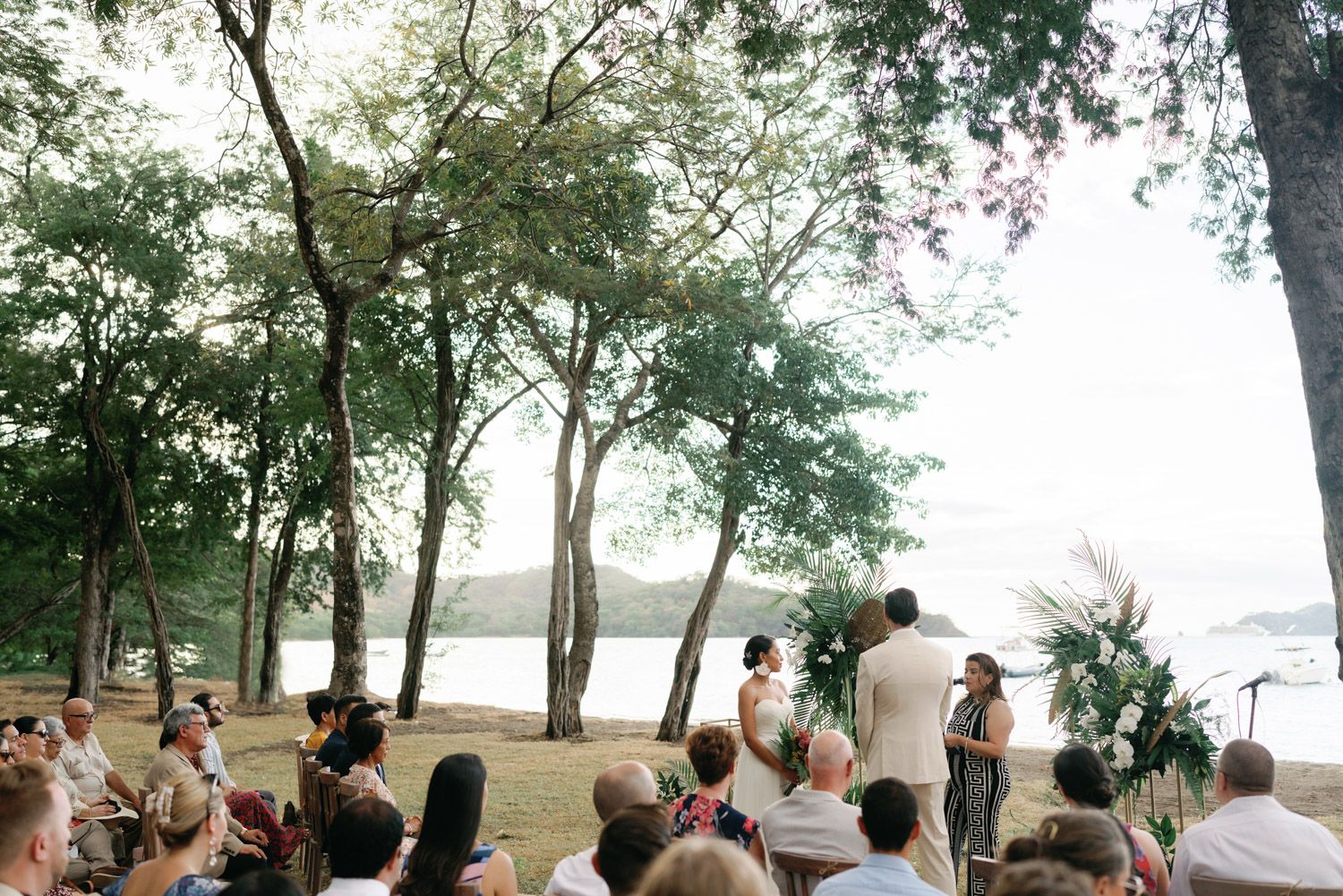 Outdoor wedding ceremony under tall trees by a beach, with guests seated watching couple exchange vows.