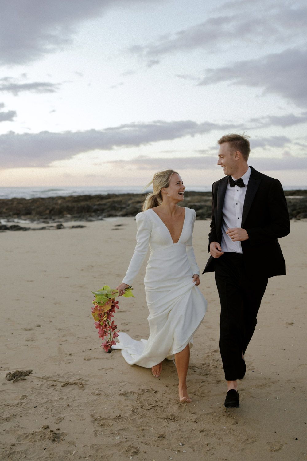 Bride in a white long-sleeve gown holding a colorful bouquet walks barefoot on the beach with a groom in a black tuxedo at sunset.