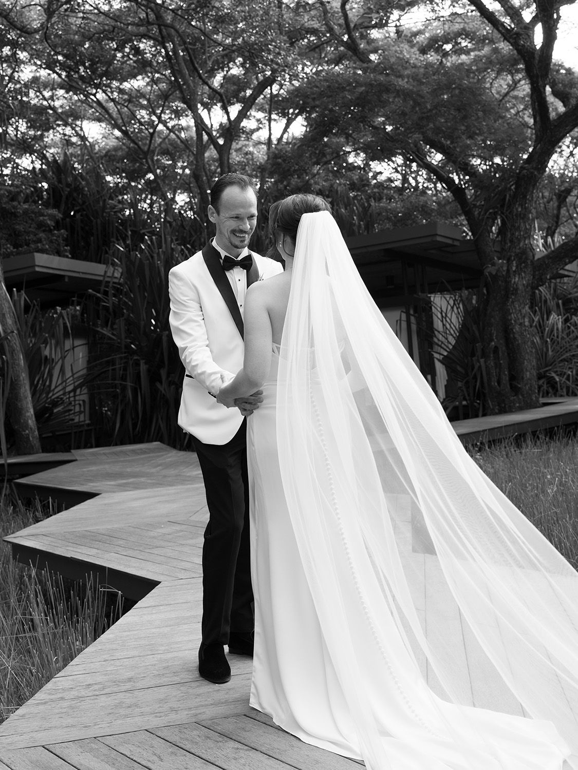 Black-and-white photograph of a groom smiling after first look at his bride on a wooden walkway surrounded by tropical trees in Costa Rica