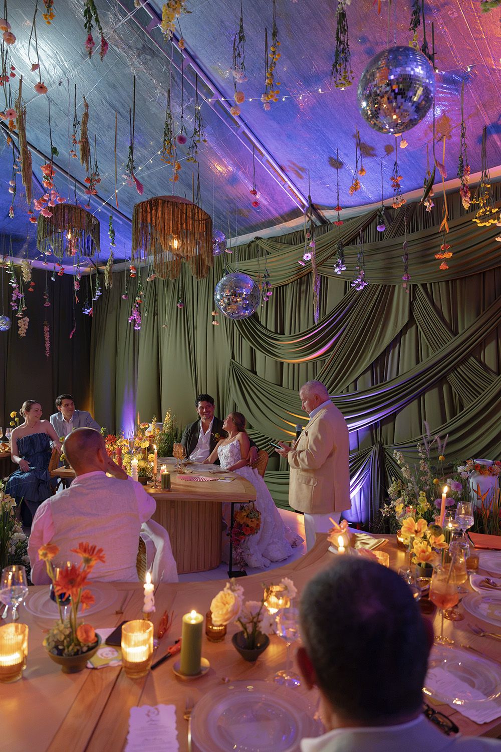 Bride and groom listening to a wedding speech at a candlelit reception in Costa Rica, surrounded by hanging flowers and green drapery.