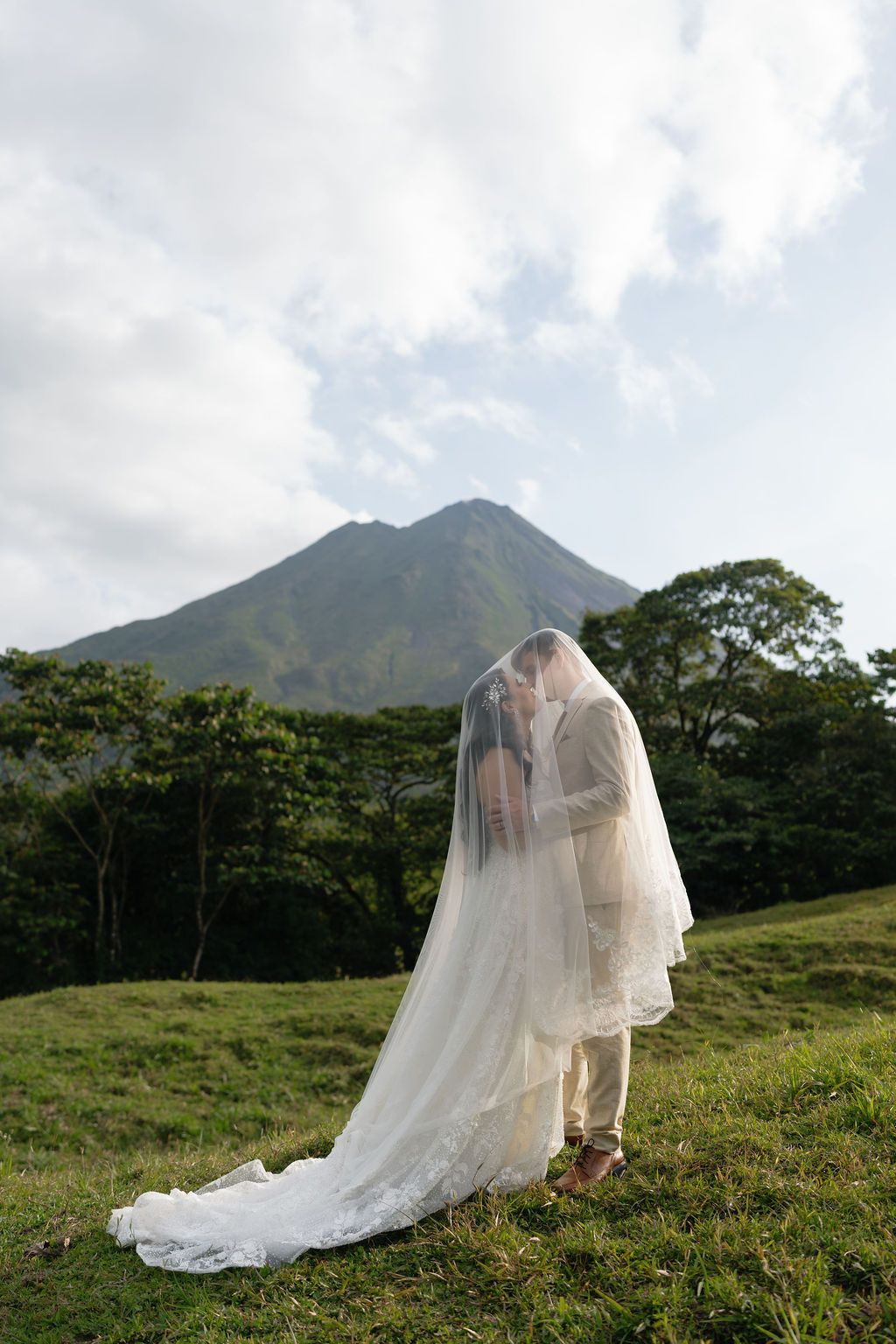 Newlyweds embracing beneath the bride’s veil with Arenal Volcano behind them