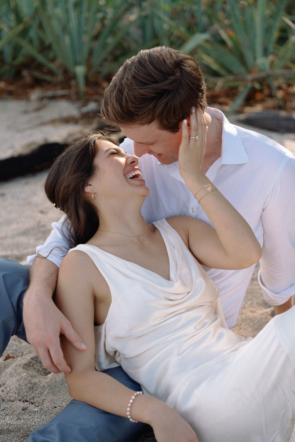 A laughing couple in white outfits embracing on a sandy beach, with a woman in a silk dress resting against a man's chest.