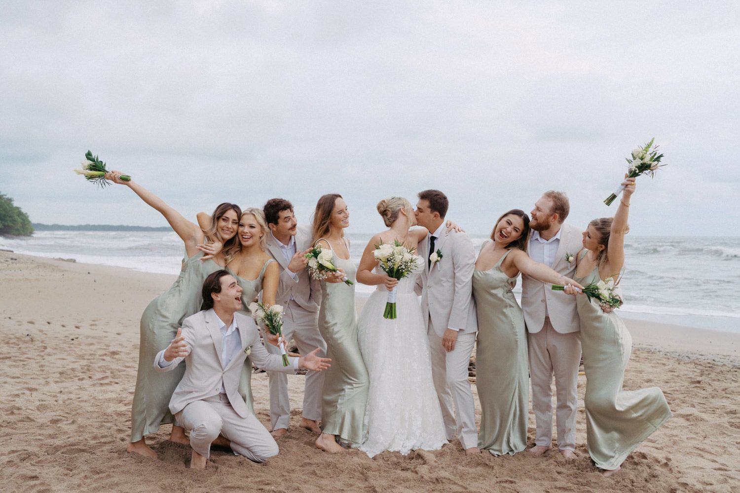 Bride and groom pose on a beach with joyful bridesmaids in sage green dresses and groomsmen in beige suits cheering with bouquets.