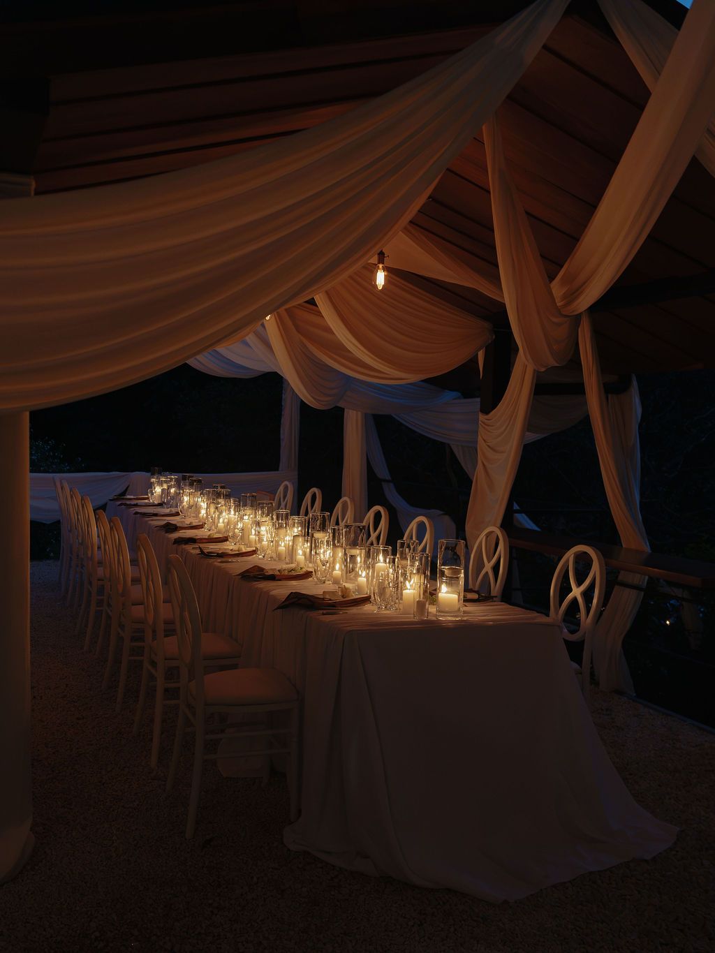 Long wedding dinner table illuminated by candles beneath flowing white drapery