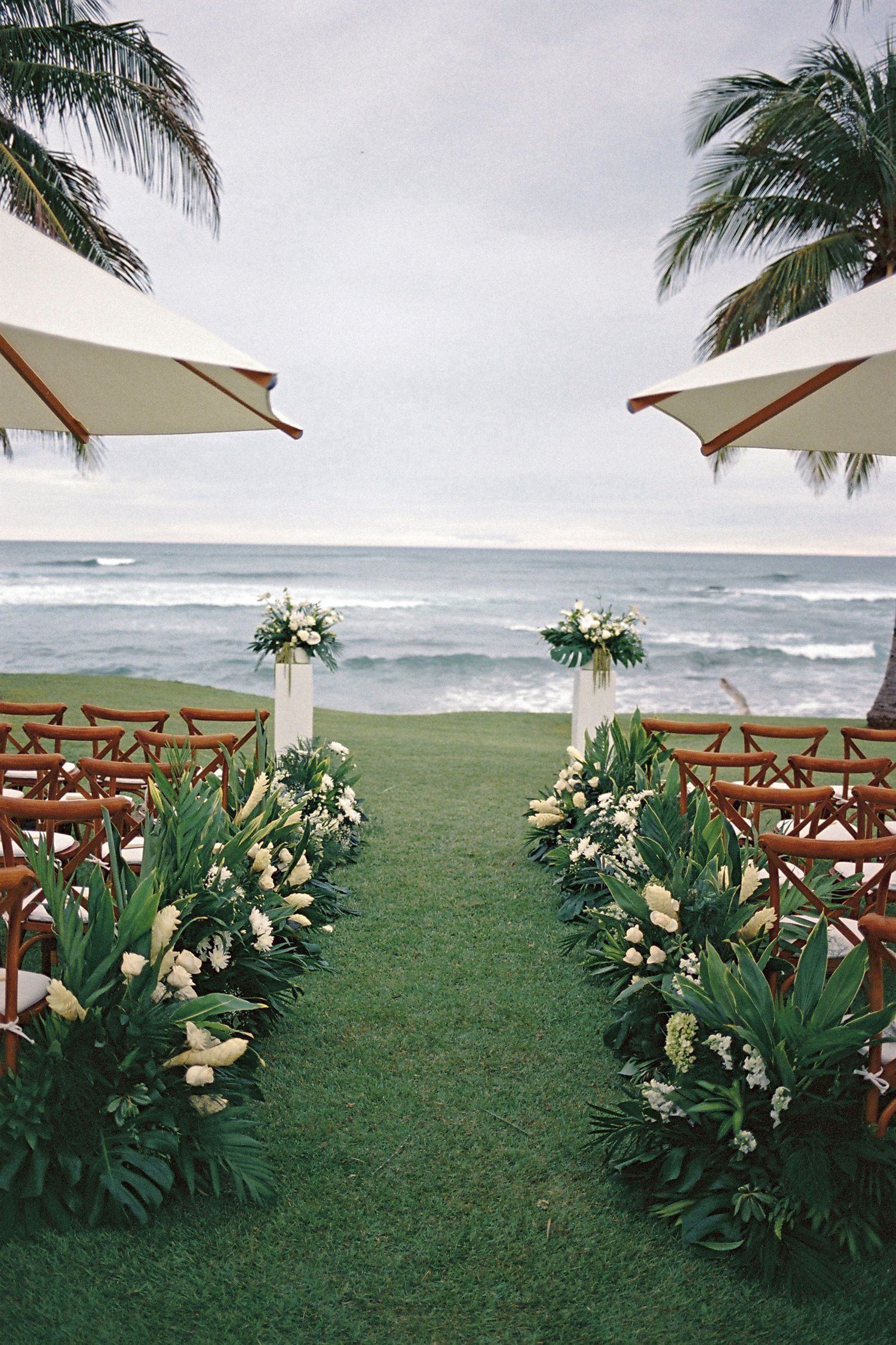 Elegant beachfront wedding aisle with tropical greenery, white floral arrangements, wooden chairs, and ocean backdrop.