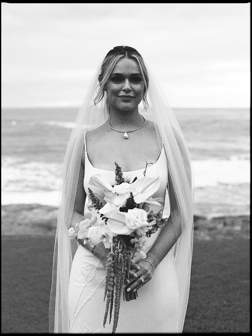 Black and white film portrait of the bride holding her bouquet by the ocean