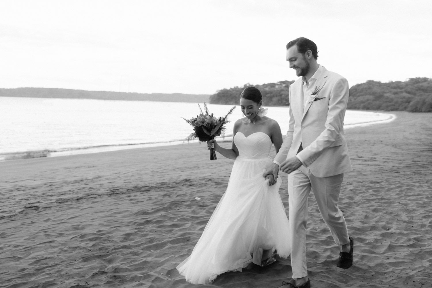 Bride and groom walking hand in hand along Playa Conchal in Costa Rica, photographed in black and white.