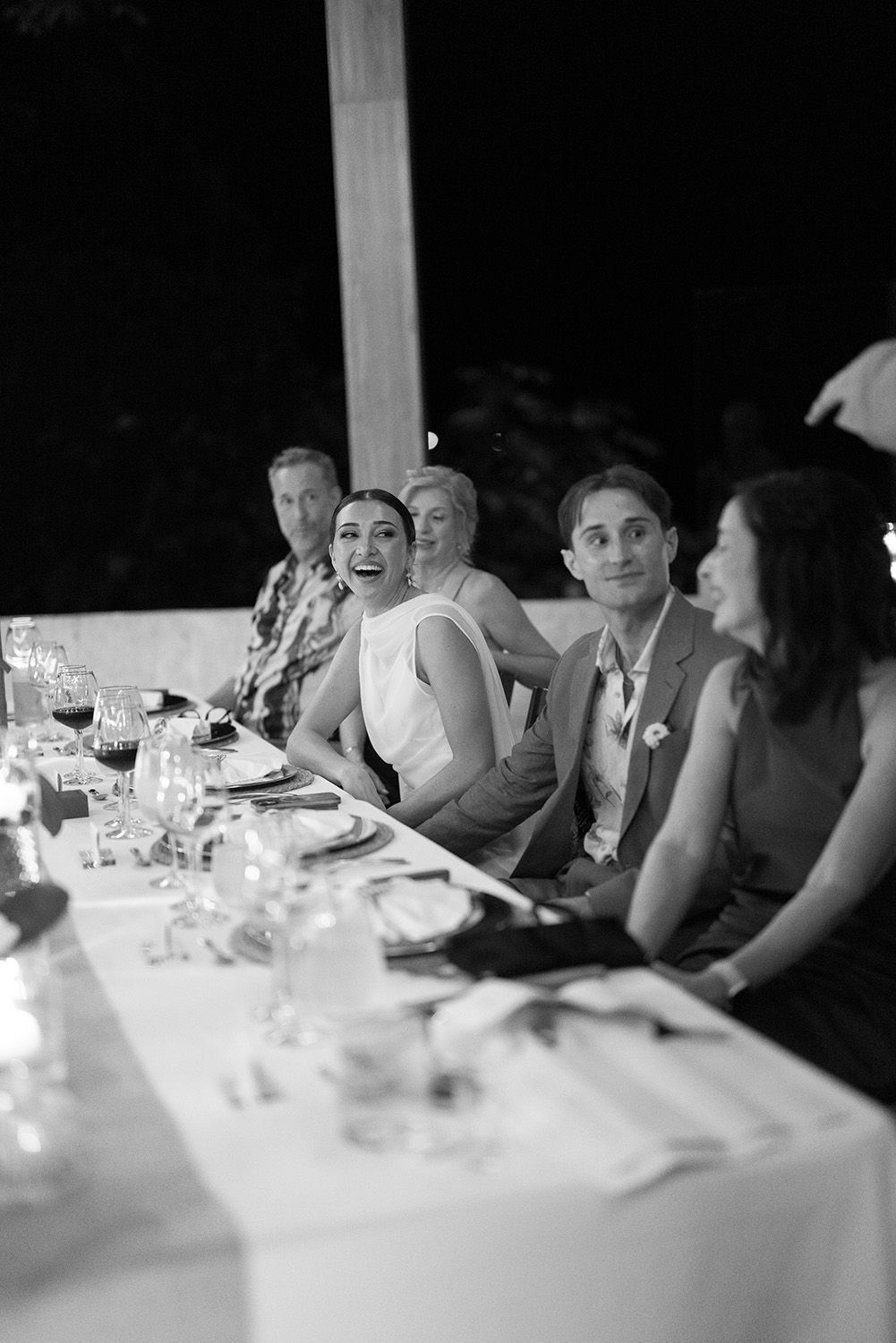 Black and white photo of wedding guests laughing and smiling at a candlelit reception dinner table.