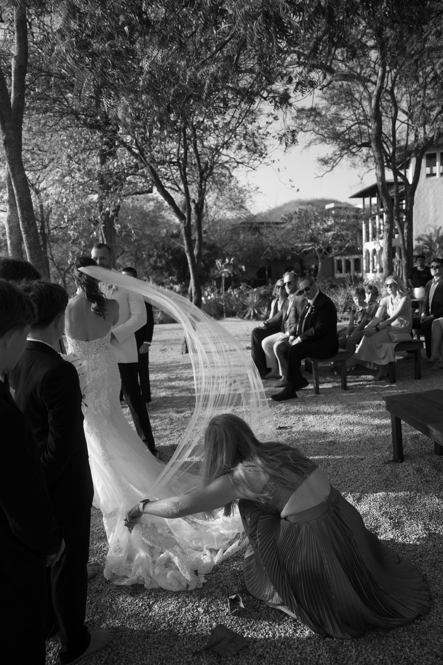 Black and white wedding photo of a bridesmaid kneeling to arrange the bride's long flowing veil as guests watch outdoors under trees.
