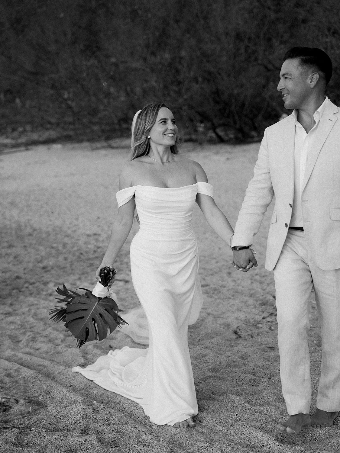Black-and-white portrait of a bride and groom smiling and holding hands while walking barefoot on a beach in Costa Rica.