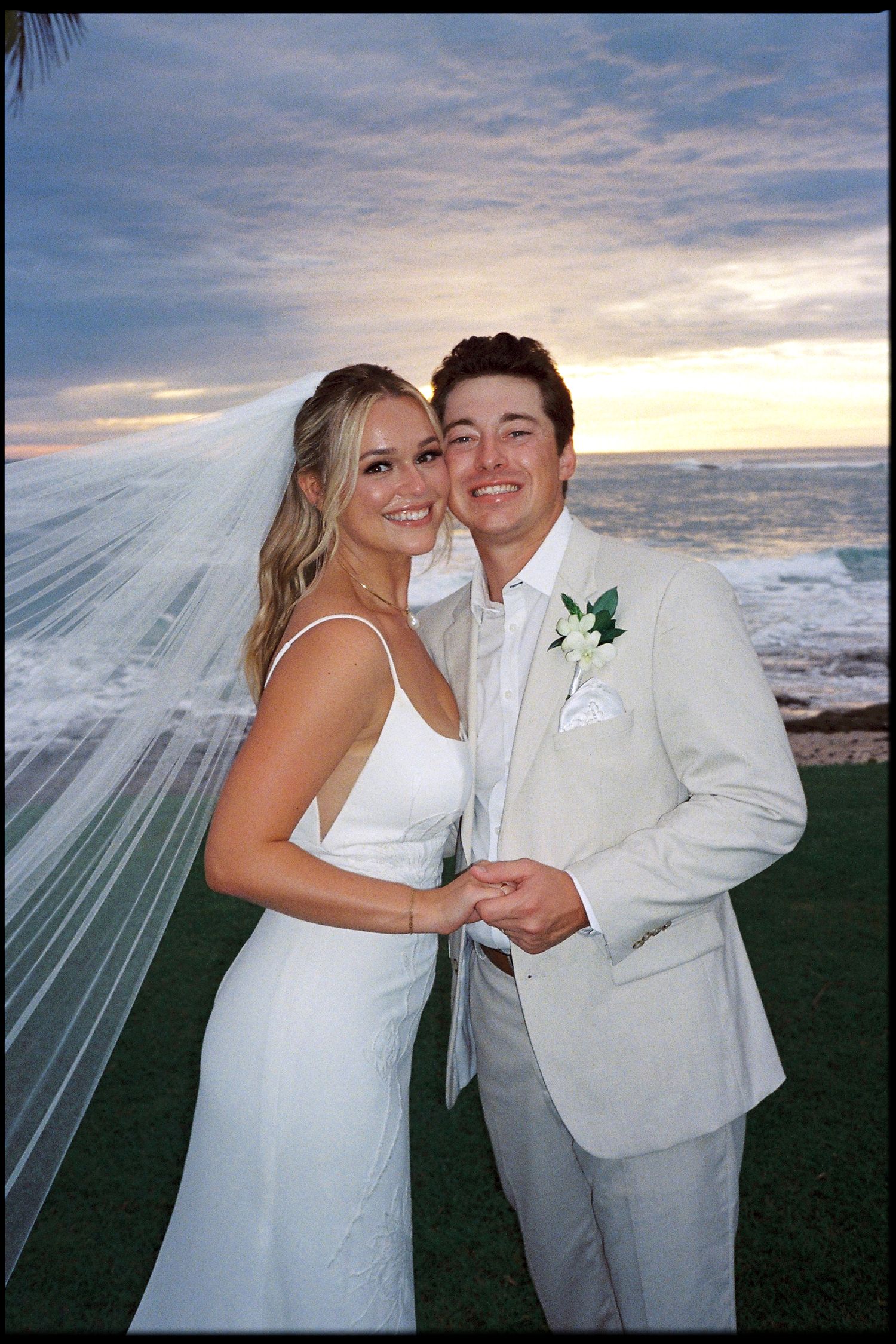 Bride and groom pose together at beachside sunset wedding, she in white gown with veil, he in cream suit with boutonniere.