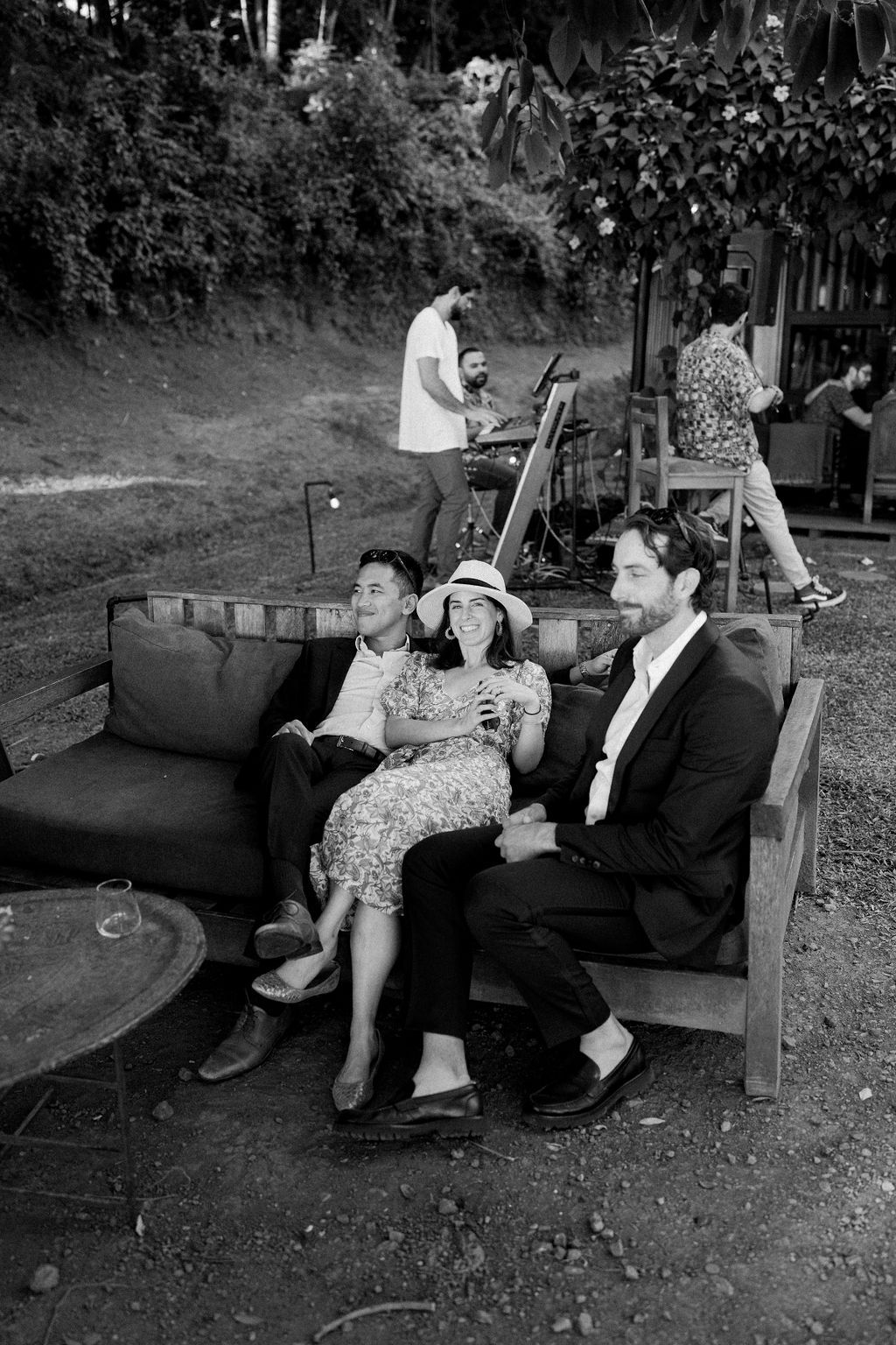 Black and white photo of three people relaxing on an outdoor wooden sofa while a musician sets up equipment in a leafy garden.