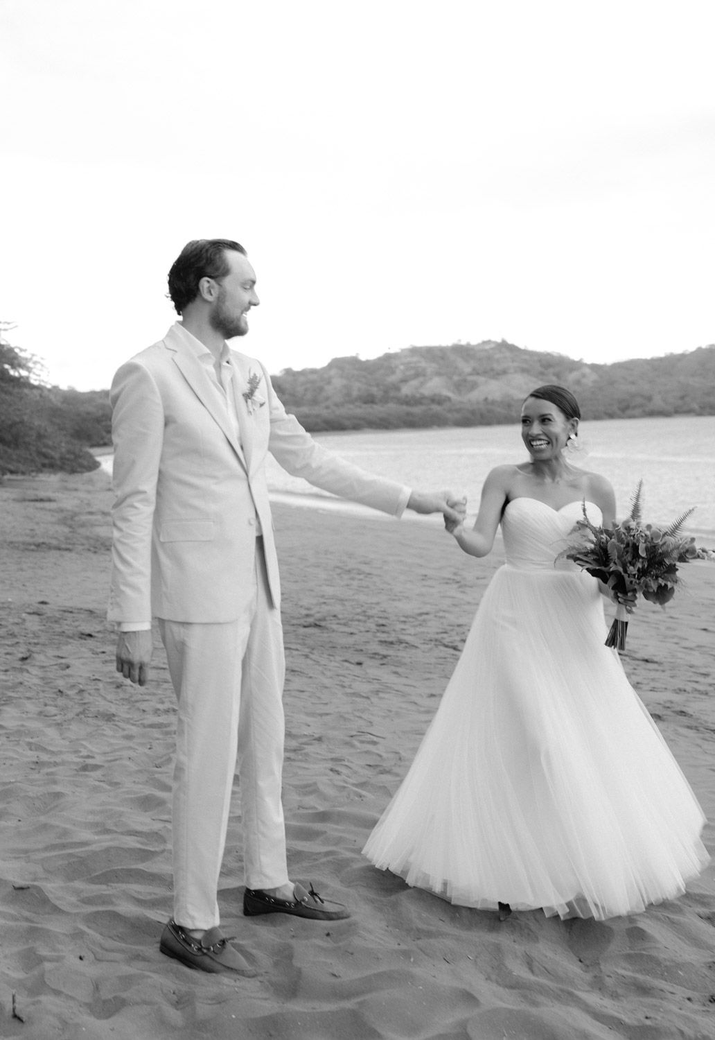 Bride in a strapless tulle gown holding a bouquet smiles and holds hands with a groom in a light suit on a beach wedding day.