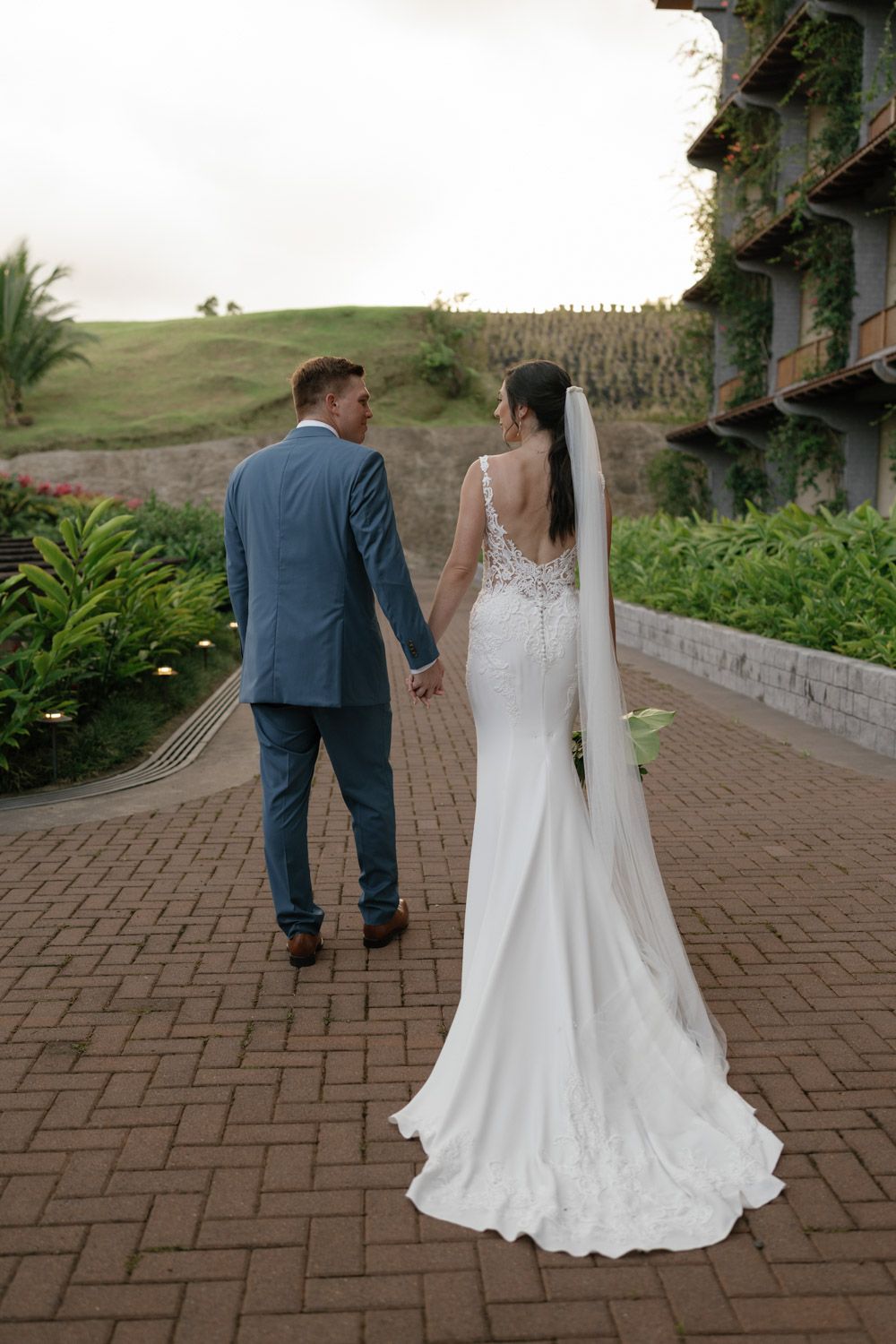 Bride in white gown with veil and groom in blue suit hold hands walking on brick path at outdoor wedding venue in Costa Rica