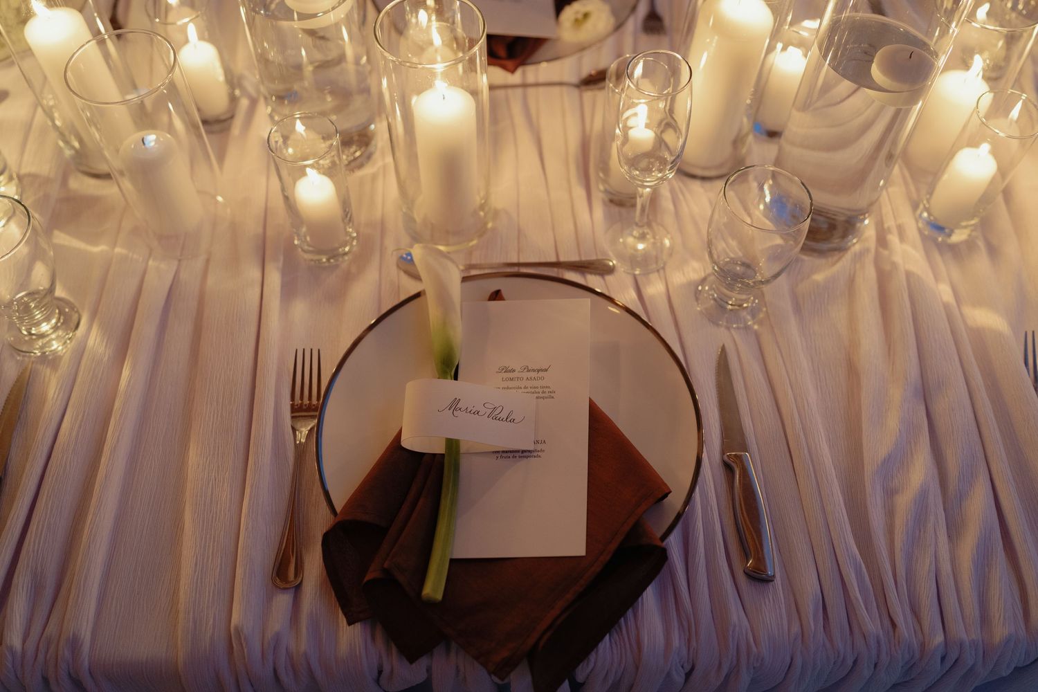 Elegant wedding reception place setting with white candles, crystal glasses, brown napkin, and personalized menu card.
