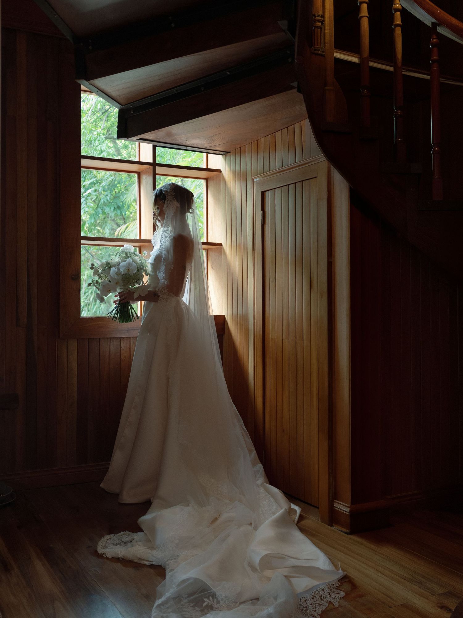 Bride in white wedding gown and veil stands by wooden staircase window holding bouquet in rustic venue.