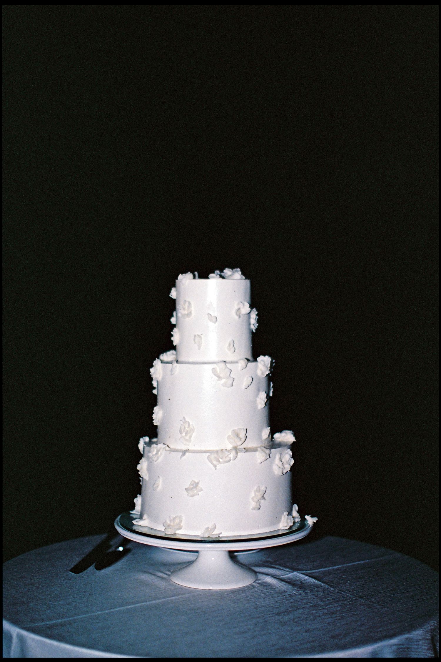 Elegant three-tier white wedding cake with floral decorations on a white pedestal stand against dark background.
