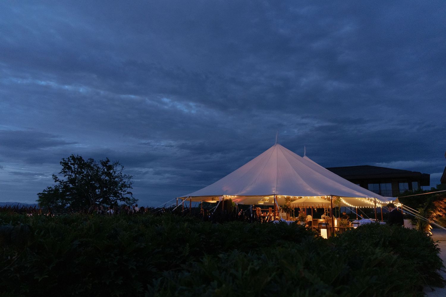Elegant white sailcloth tent glowing at dusk under dramatic cloudy sky at an outdoor evening event.