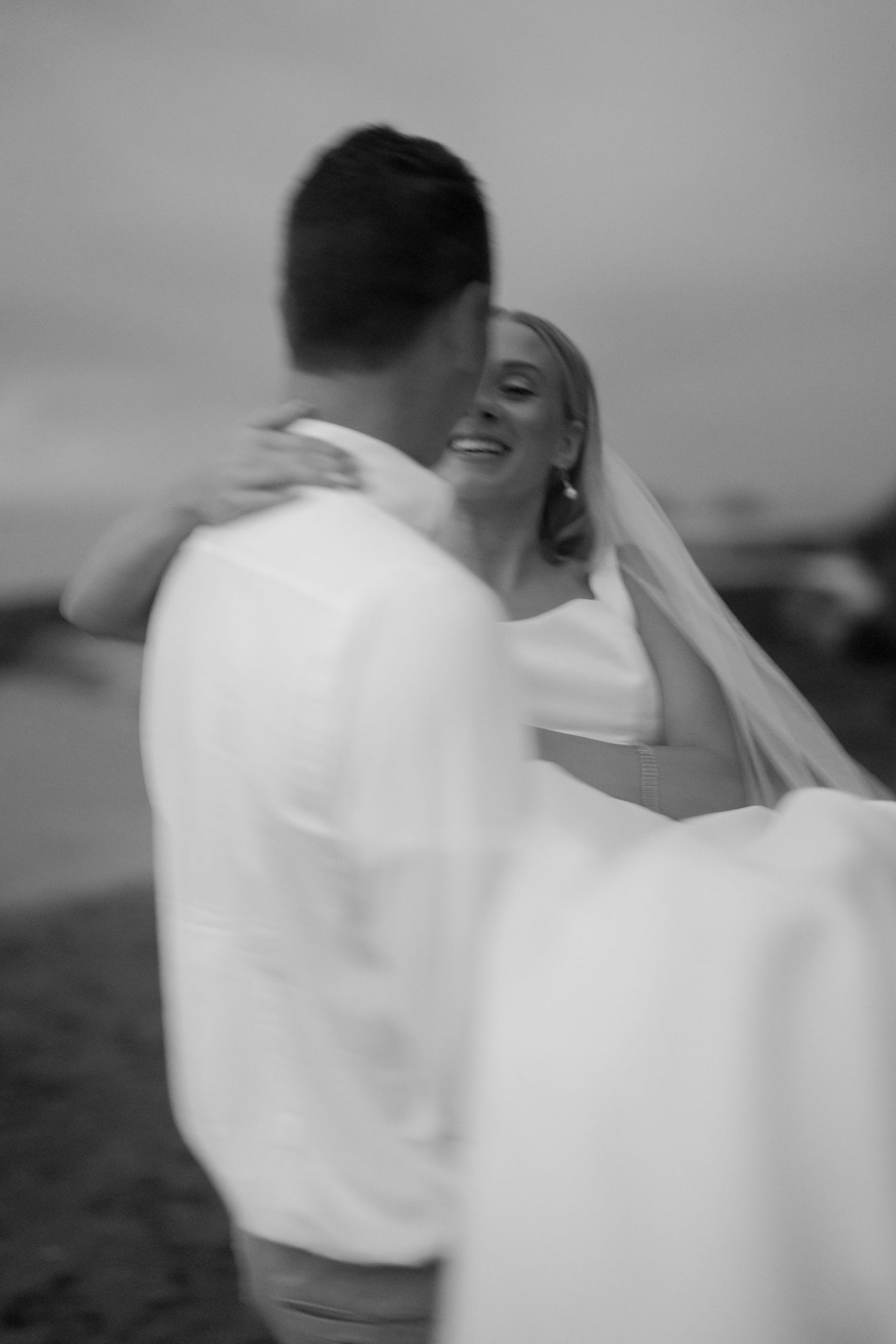 Black and white photo of a smiling bride in a veil being lifted in the arms of her groom in a white shirt at a seaside wedding.