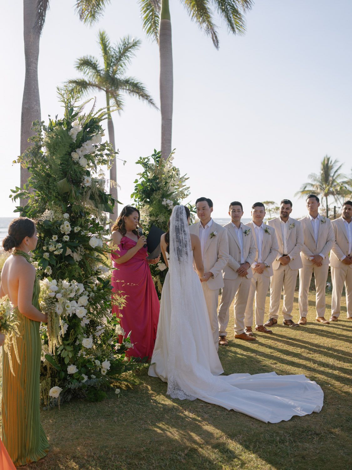 Outdoor beach wedding ceremony with bride, groom, groomsmen in tan suits, and floral arch surrounded by palm trees in Costa Rica