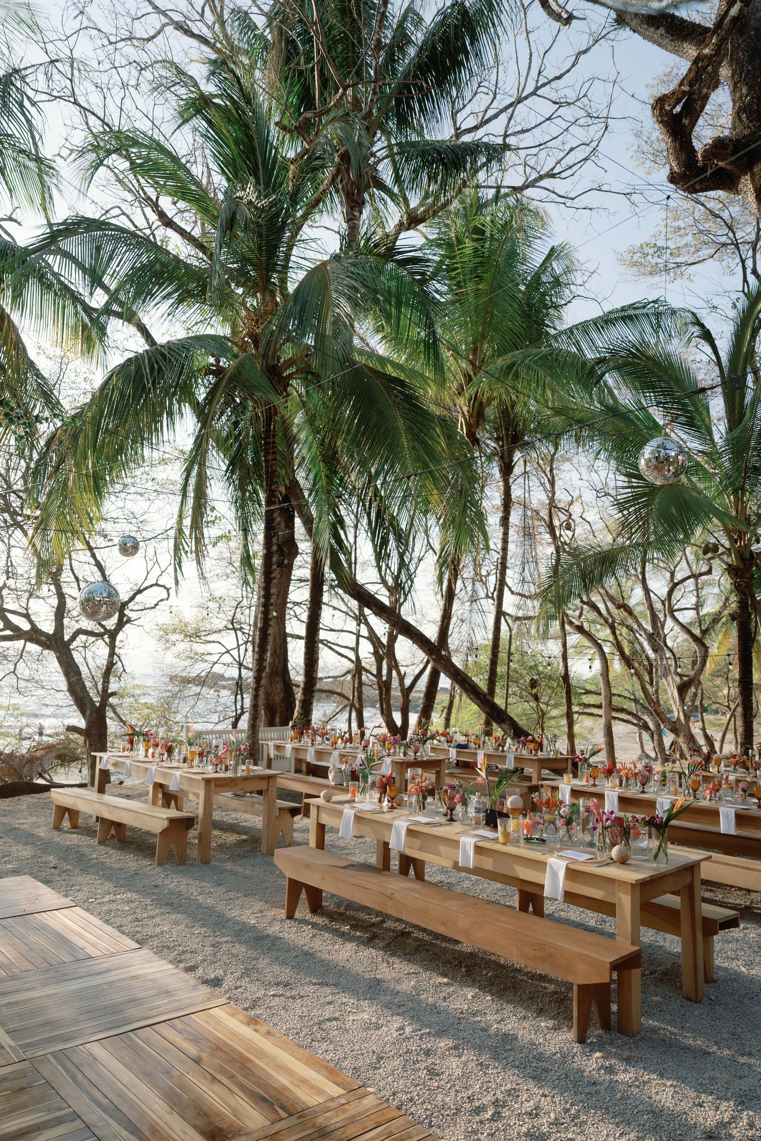 Outdoor wedding reception with wooden tables and benches set under tropical palm trees on a gravel surface.