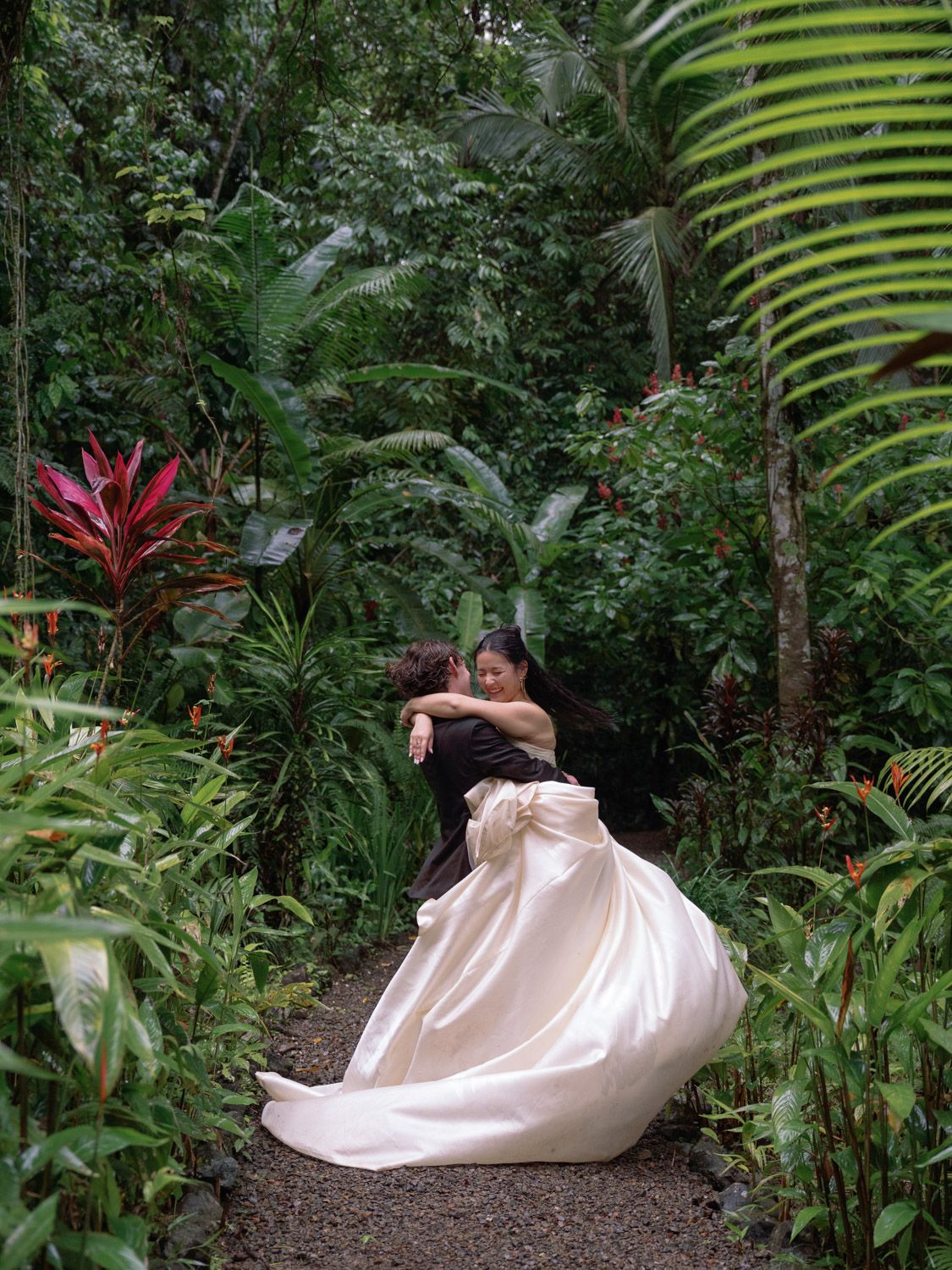 Couple embracing on a lush tropical garden path, bride in flowing white gown, surrounded by vibrant greenery in Costa Rica