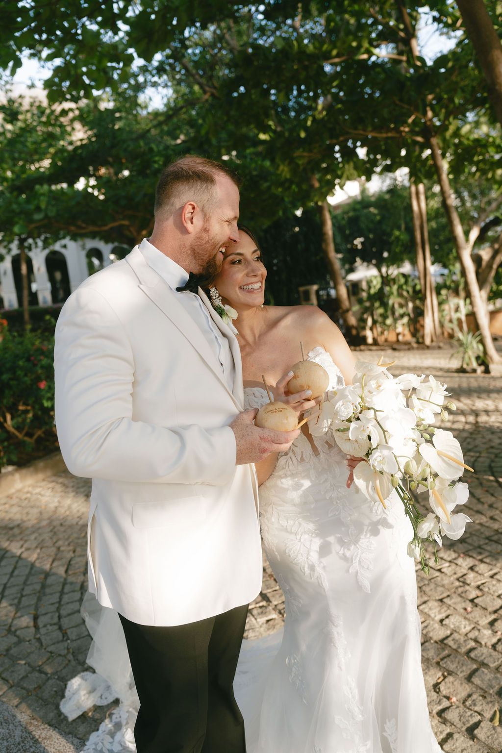 Bride in a white lace gown and groom in a white tuxedo jacket embrace outdoors, holding coconuts and a bouquet of white flowers.