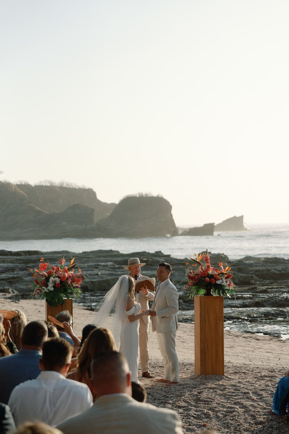 Couple exchanges vows at a romantic beach wedding ceremony with tropical floral arrangements and rocky ocean backdrop in costa rica