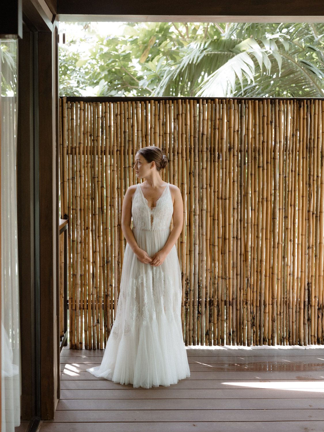 Bride in a flowing white lace wedding gown stands on a wooden deck beside a bamboo fence with tropical palm leaves behind her.