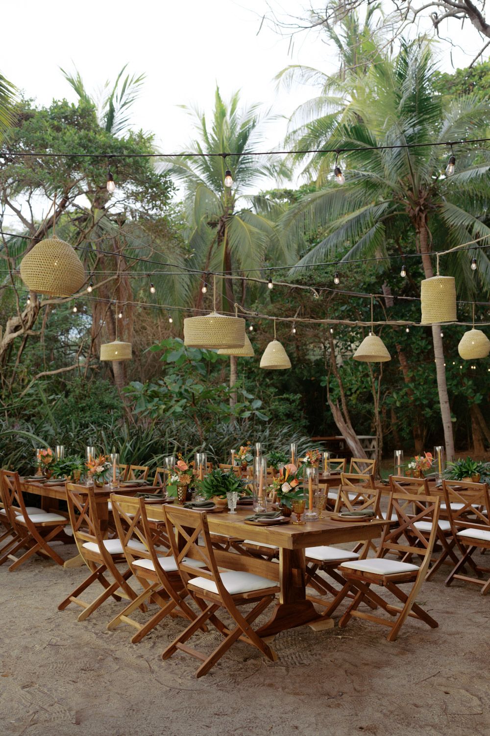Rustic wooden tables with folding chairs and woven pendant lamps set for an outdoor tropical beach wedding reception under palm trees.