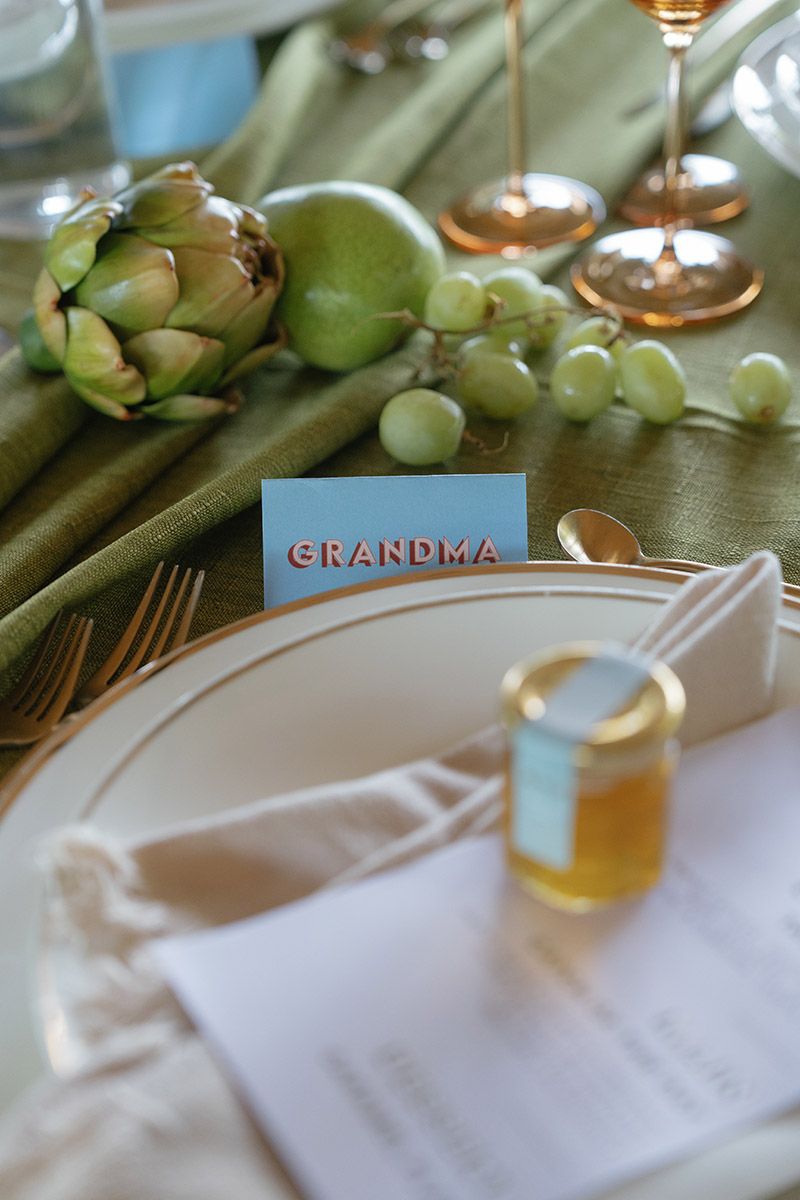 Elegant wedding table setting with a Grandma place card, artichoke centerpiece, grapes, honey jar, and gold flatware.