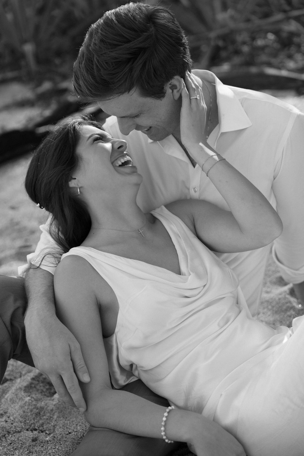 Black and white photo of a laughing woman in a white dress reclining on a man's lap as he smiles down at her on a beach.