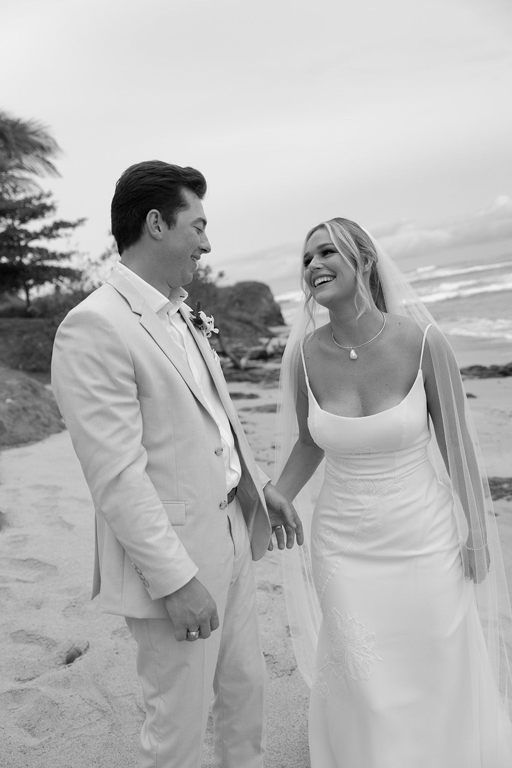 Black-and-white portrait of a bride and groom laughing together on a beach in Costa Rica, with ocean waves behind them
