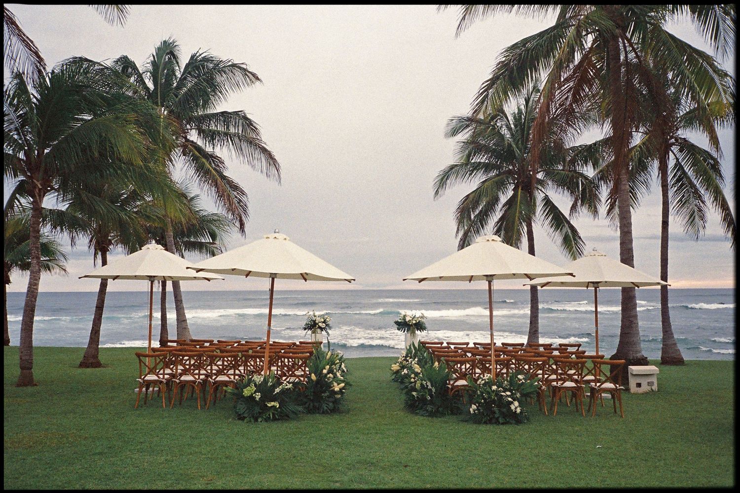 Beachside wedding setup with wooden chairs, white umbrellas, and tropical palm trees overlooking the ocean.