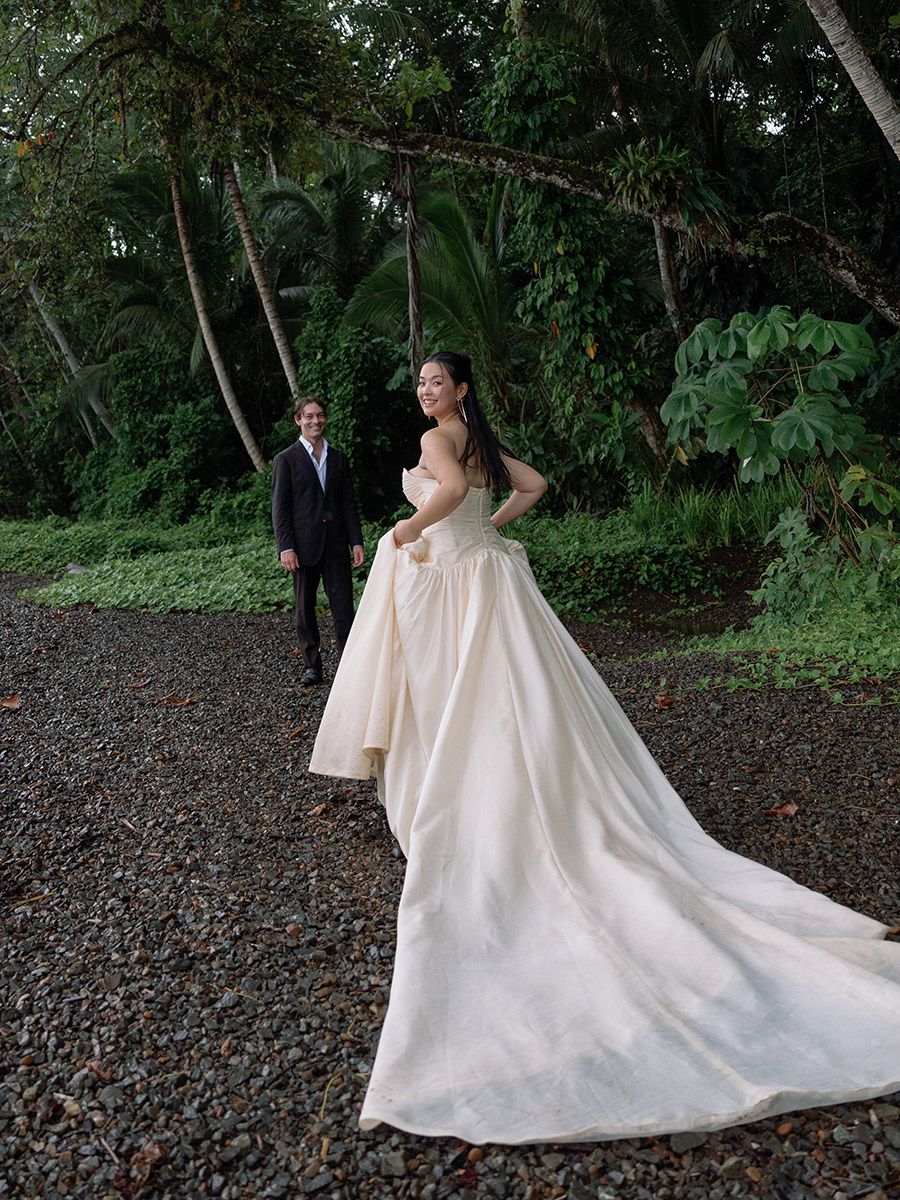 Bride in an elegant gown with a long train stands on a gravel path, groom in a suit behind her, lush tropical greenery.